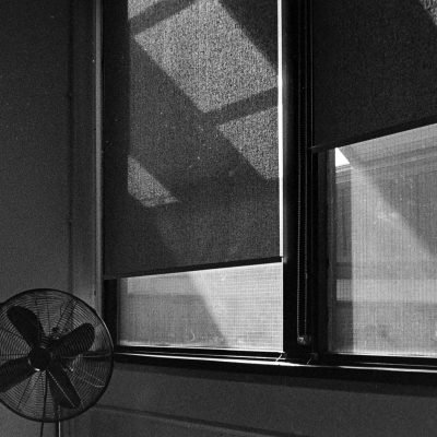 Black and white photo depicting a vintage fan next to shadowed windows, creating a moody interior scene.